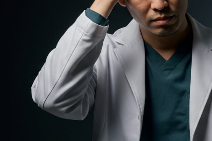 医療用スクラブと白衣を着た男性医師が、暗い背景の中で額に手を当て、疲れた表情を見せている様子