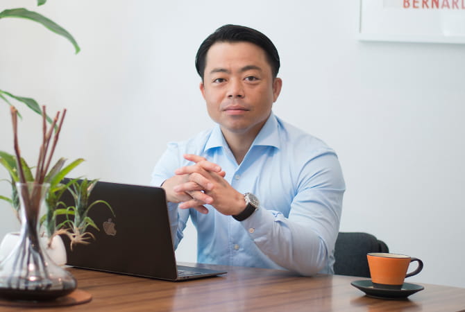 ノートパソコンの前で手を組みながらカメラ目線で座る代表の写真